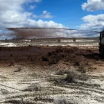 A truck sprays a stream of dark liquid or soil across a barren, dry landscape under a partly cloudy sky, with two workers standing on top of the vehicle.