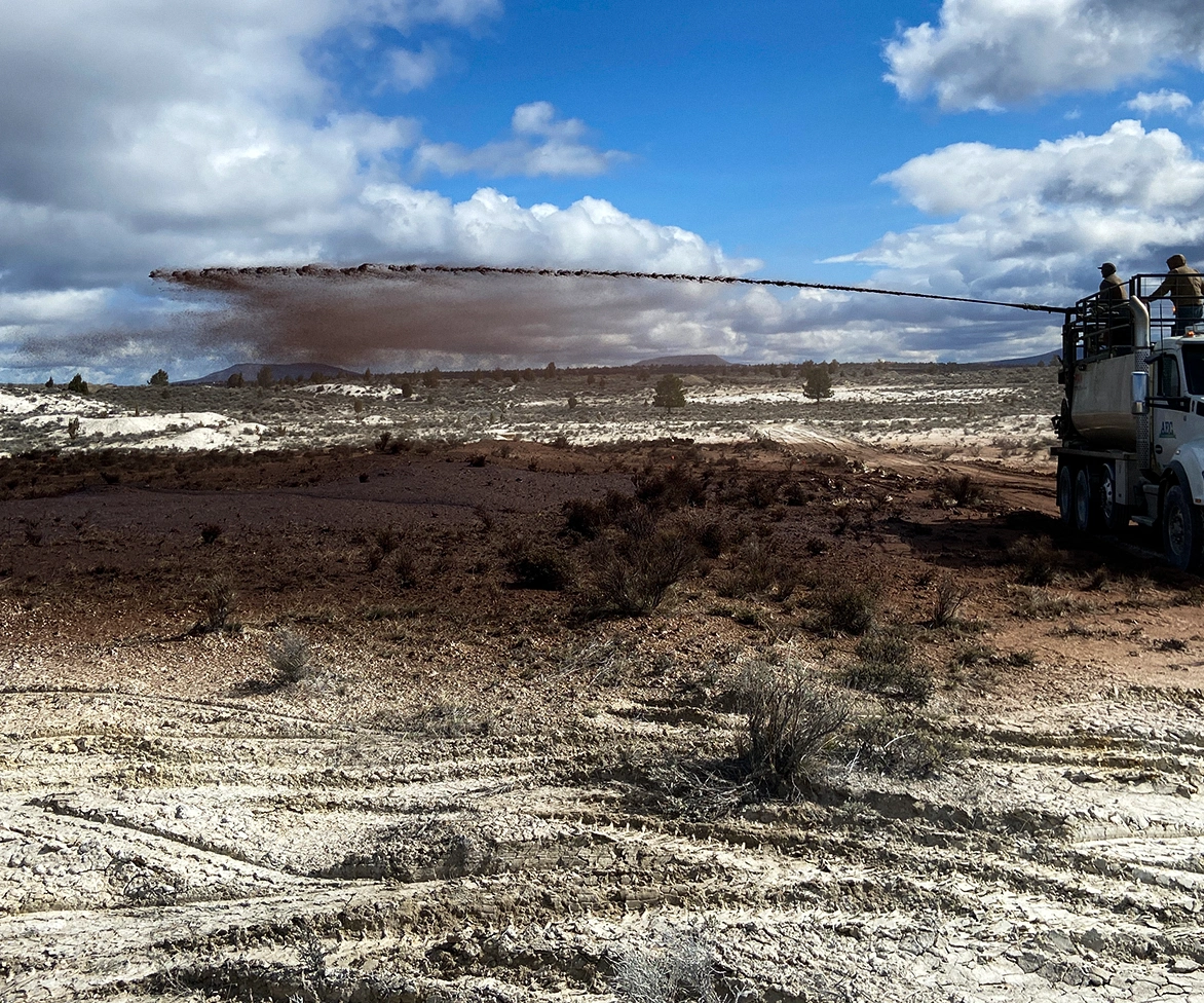 A truck sprays a stream of dark liquid or soil across a barren, dry landscape under a partly cloudy sky, with two workers standing on top of the vehicle.