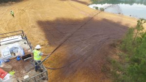 A worker in a safety vest and helmet sprays a substance onto a sloped, bare patch of land near a pond, while another worker stands in the distance. The area appears to be part of a construction or land restoration site.