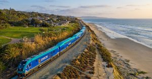 A blue and silver train travels along a coastal railway with the ocean on one side and green parkland with houses on the other under a partly cloudy sky at sunset.