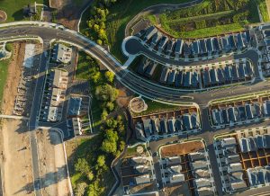 Aerial view of a suburban neighborhood with houses, streets, sidewalks, green areas, and some ongoing construction in one section. Trees and landscaped yards are visible throughout the community.