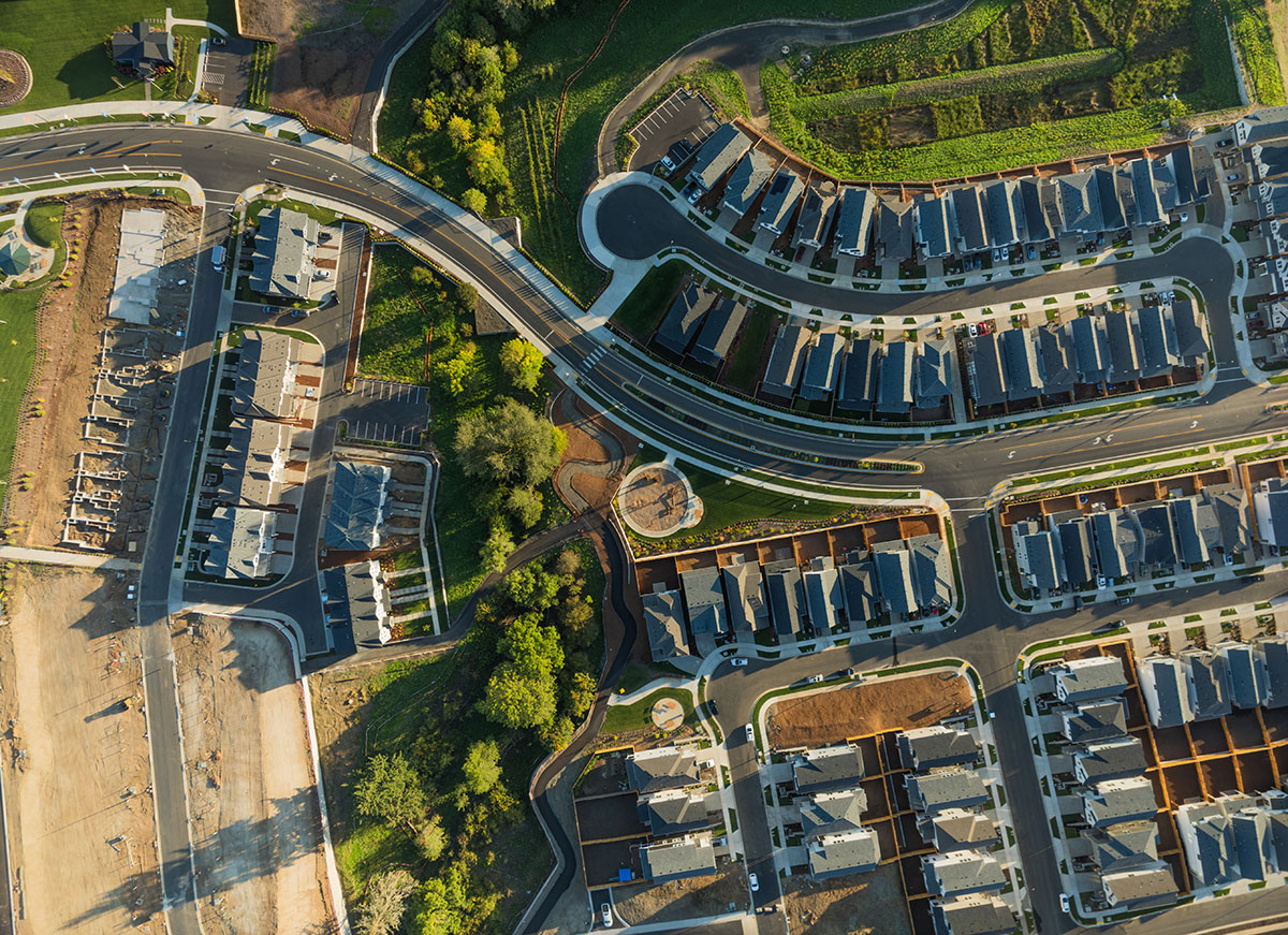 Aerial view of a suburban neighborhood with houses, streets, sidewalks, green areas, and some ongoing construction in one section. Trees and landscaped yards are visible throughout the community.