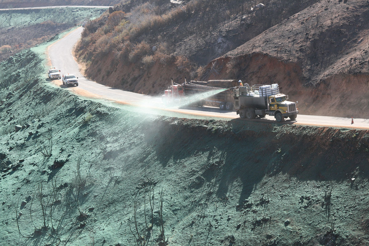 A truck sprays a green substance on a hillside next to a road, likely for erosion control or re-vegetation, while other vehicles drive nearby. The area shows signs of previous fire damage.
