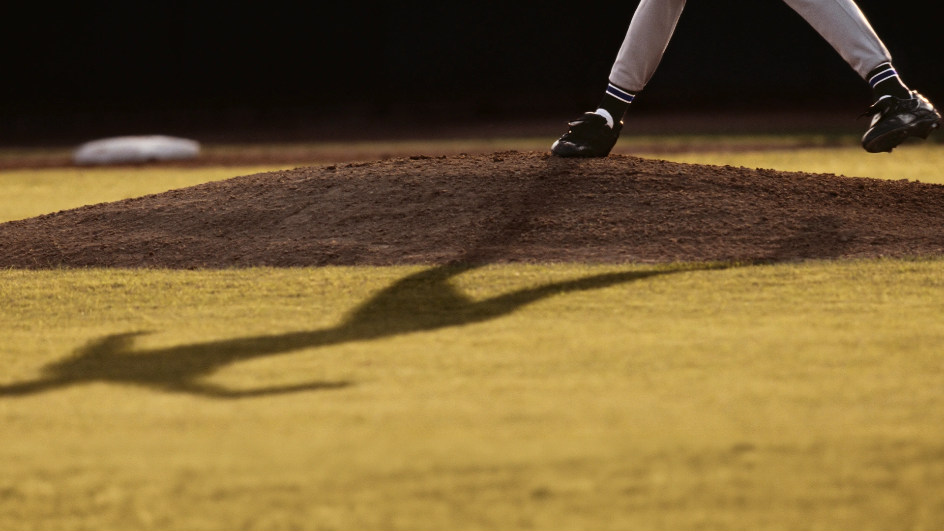 A baseball pitchers legs and shadow are visible on a pitchers mound, mid-pitch, with the field in the background.