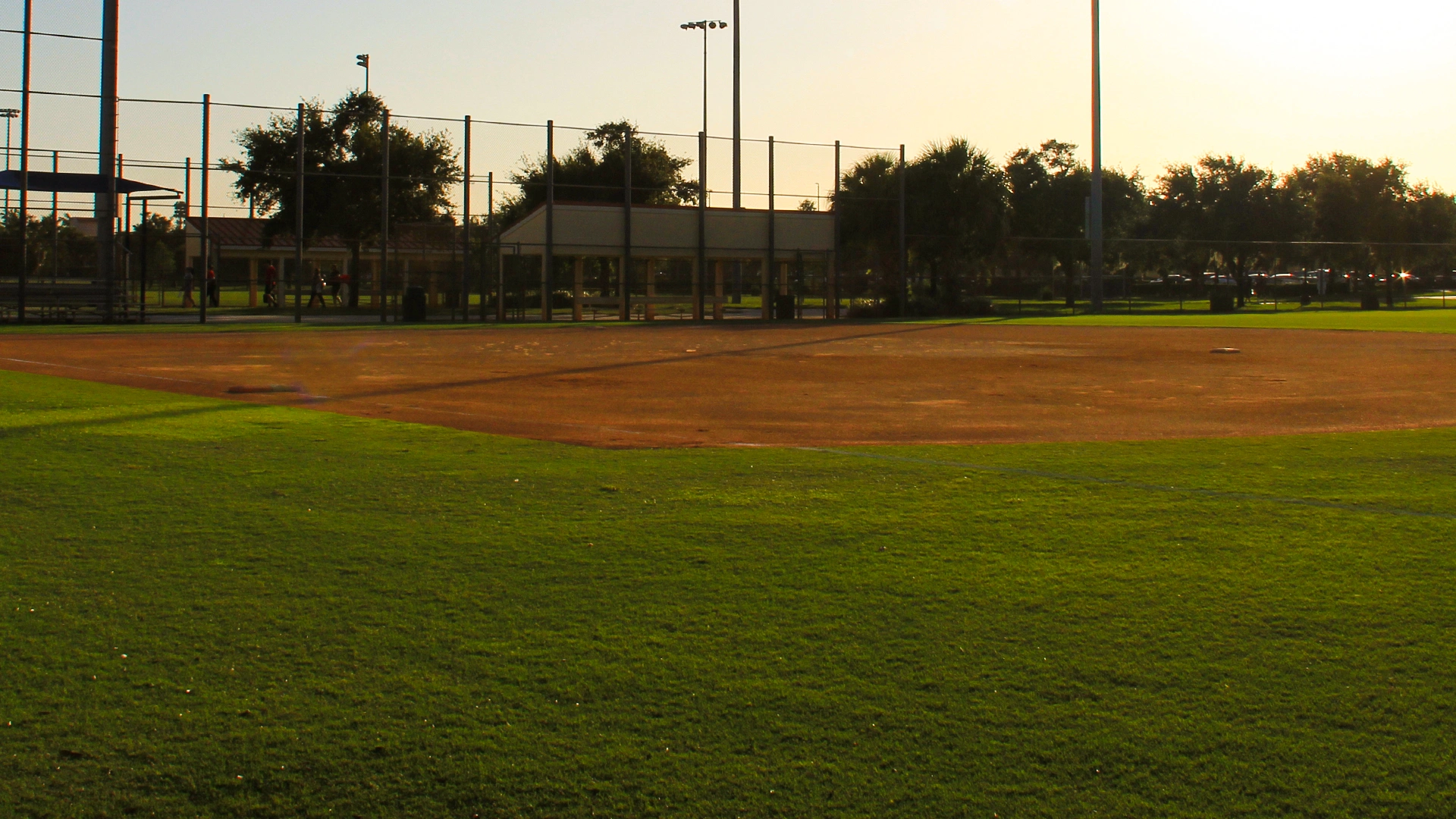 Baseball field at sunset with green grass, dirt infield, tall fence, trees, and a building in the background. Shadows and warm light cover the scene.