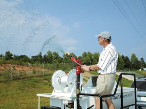 A person in a cap and striped shirt sprays green hydroseeding mixture onto a grassy slope using a large hose, with trees and a fence in the background.