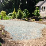 A yard with bare soil and scattered plants surrounds a patch of blue-green gravel in front of a house, with trees and greenery in the background.