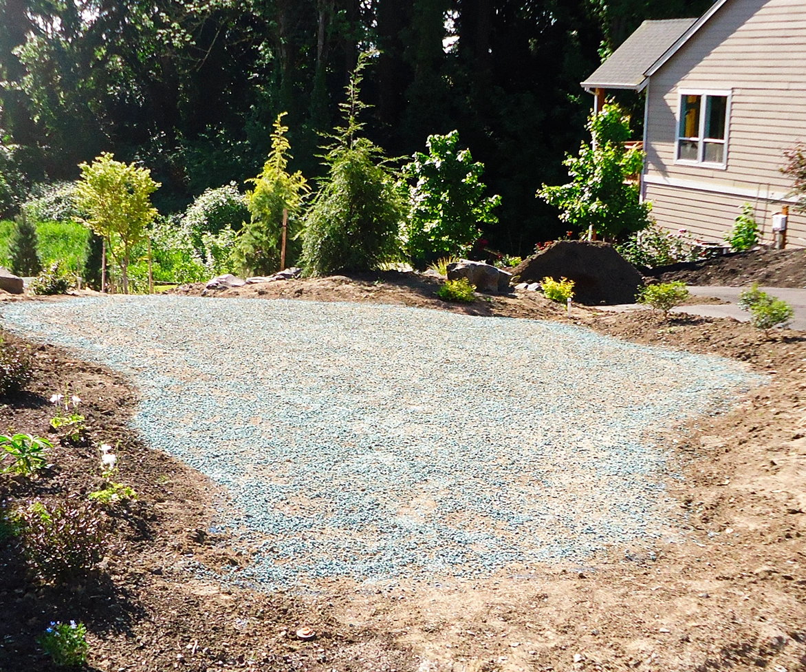 A yard with bare soil and scattered plants surrounds a patch of blue-green gravel in front of a house, with trees and greenery in the background.