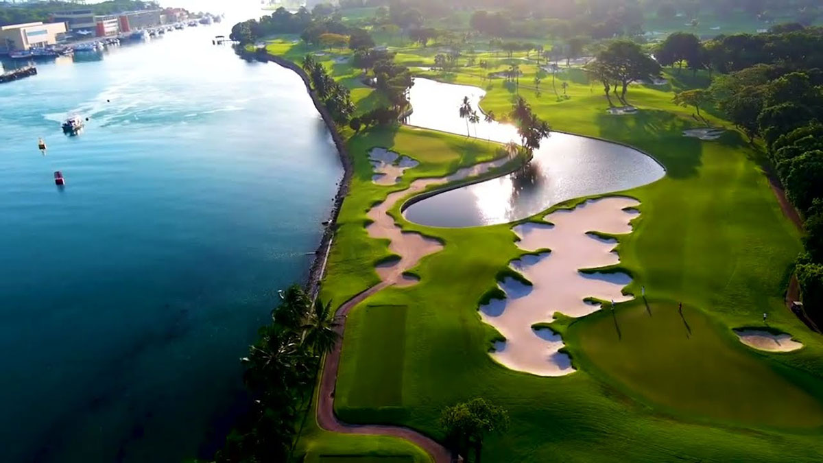 Aerial view of a scenic golf course next to a body of water, with sand traps, green fairways, trees, and a pond reflecting sunlight.