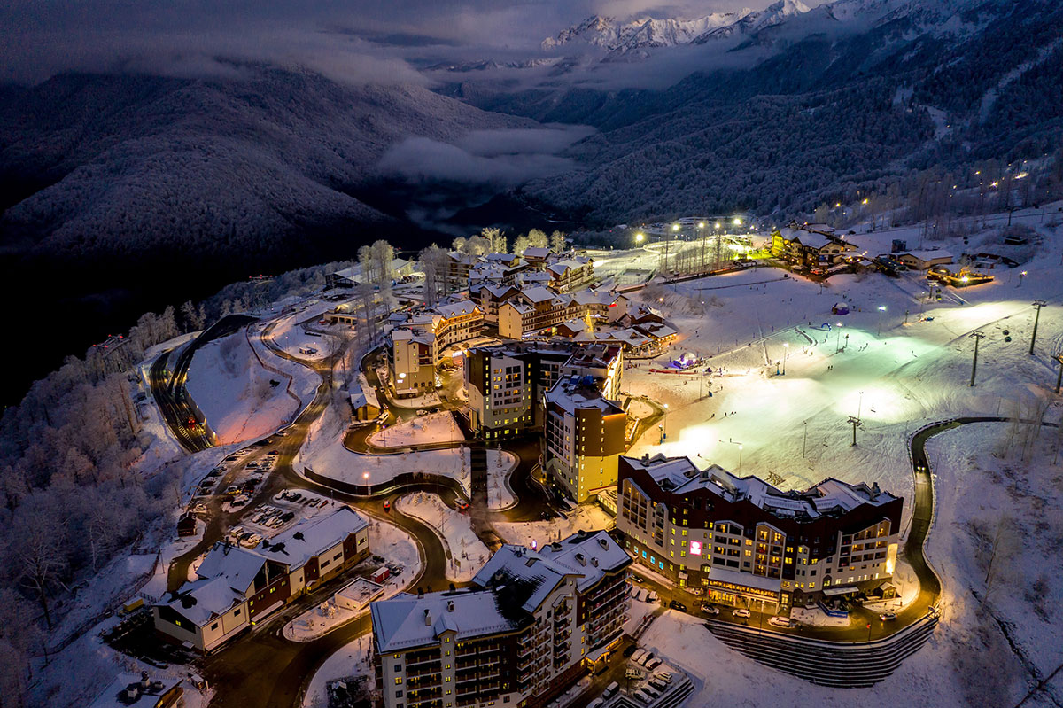Aerial view of a snow-covered mountain resort at night, with illuminated buildings and winding roads surrounded by trees and mountains under a cloudy sky.