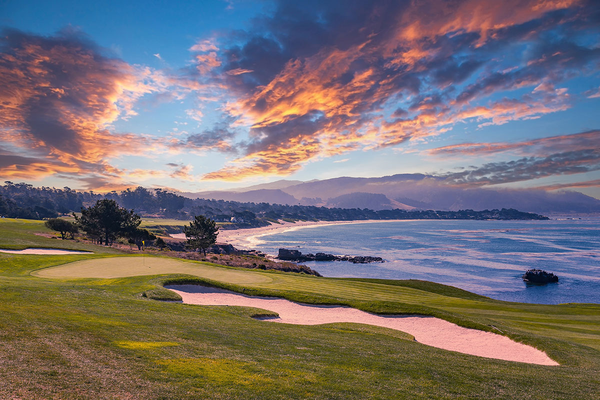 A scenic golf course by the ocean at sunset, with sand bunkers in the foreground, rolling green hills, and dramatic clouds in the colorful sky.