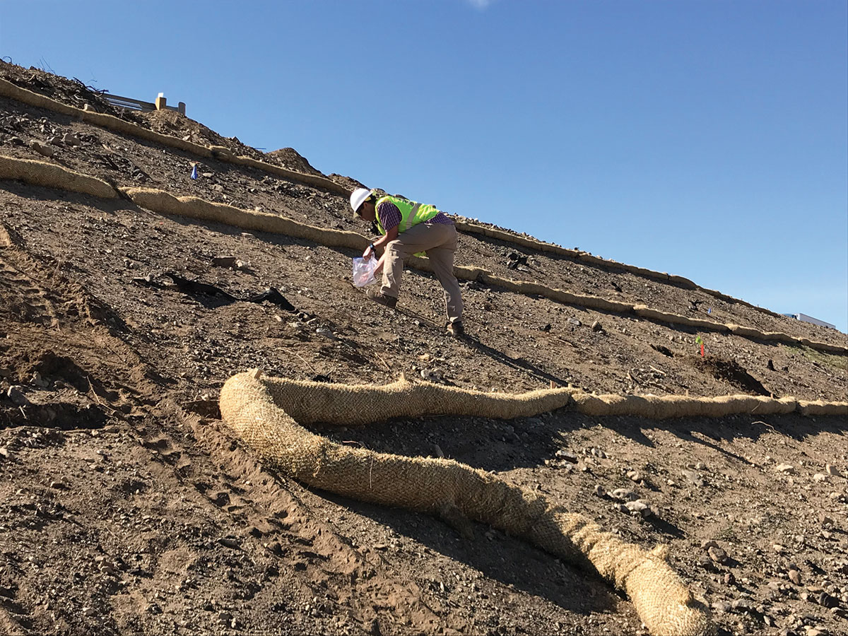 A worker in a safety vest and helmet spreads material on a steep, barren slope with straw wattles for erosion control under a clear blue sky.