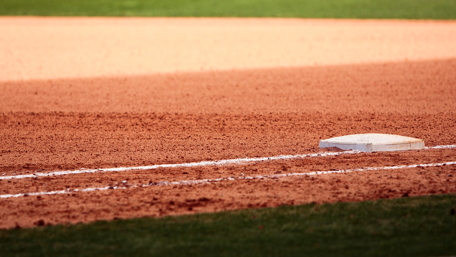 A close-up view of a baseball field showing third base and the infield dirt, with white chalk lines marking the boundaries. The grass of the outfield is visible in the background.