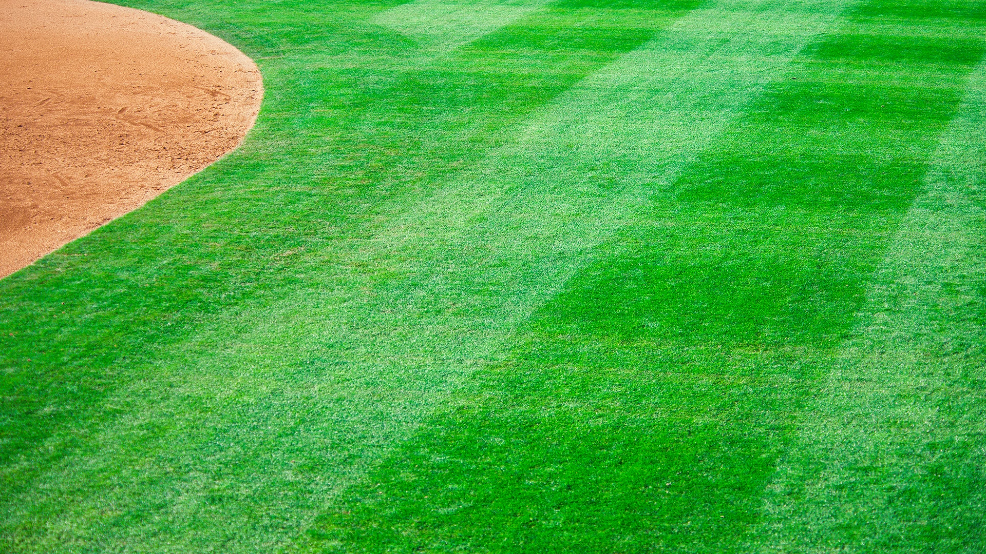 A close-up view of a baseball field, showing the edge of the dirt infield and the grassy outfield with visible mowing patterns.
