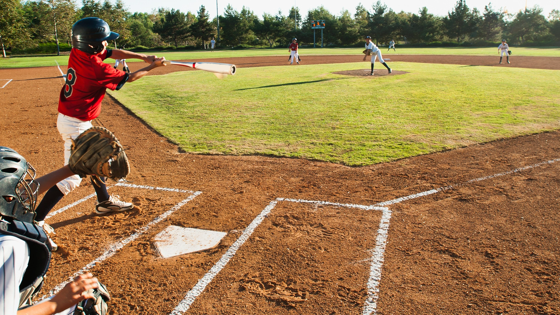 A baseball player in a red jersey swings at a pitch while the catcher crouches behind home plate; the pitcher and other fielders are visible on a sunny day.