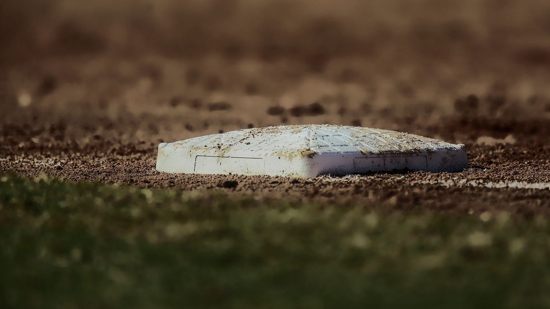 A close-up of a white baseball base on a dirt infield, with some grass visible in the foreground and blurred brown dirt in the background.