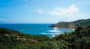 A coastal landscape with lush green hills, a winding road, and blue ocean waves meeting the rocky shore under a bright, clear sky.