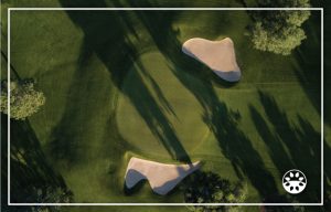 Aerial view of a golf green with two sand bunkers, long tree shadows, and a white border frame around the image.