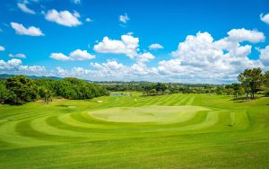 A golf course under a bright blue sky with scattered white clouds, surrounded by trees and hills. The green grass is neatly mowed with visible fairways and a flag on the putting green.