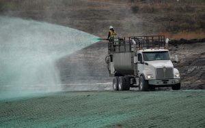 A worker stands on a large white truck spraying green hydroseed onto barren ground at a construction site, promoting grass growth and erosion control.
