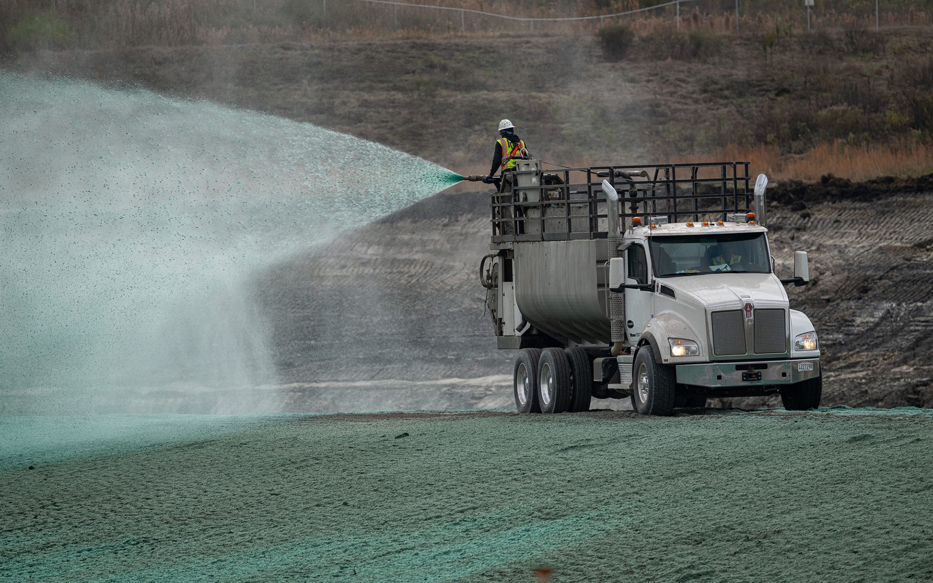 A worker stands on a large white truck spraying green hydroseed onto barren ground at a construction site, promoting grass growth and erosion control.