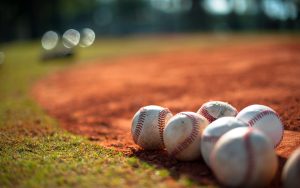 Several baseballs rest on reddish dirt near the edge of a baseball field, with grass visible in the foreground and background blurred.