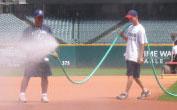 Two people stand on a baseball field; one is spraying water from a hose, and the spray partially covers the other person, who is wearing a cap and standing on dirt near green grass.