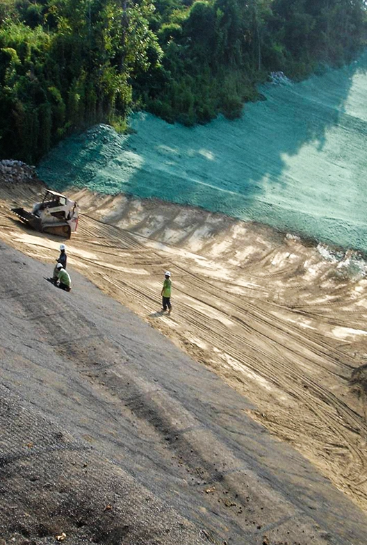 Three workers in safety vests stand on a sloped dirt embankment, while a bulldozer operates nearby. Part of the hill is covered with green erosion control material. Trees border the site.