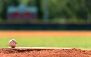 A baseball rests on the pitchers mound of a sunny baseball field, with green grass and an out-of-focus scoreboard in the background.