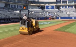A person drives a yellow steamroller on a baseball field near the infield, with empty stadium seats and a press box visible in the background.