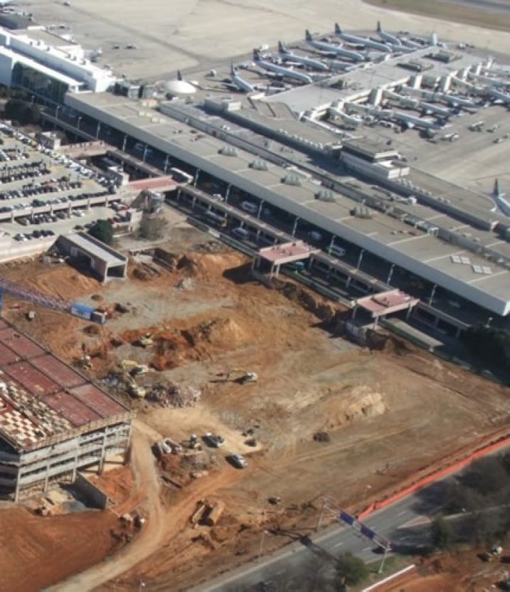 Aerial view of an airport terminal with parked airplanes; nearby, a large construction site with exposed dirt, machinery, and an unfinished building is visible.