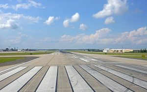 View of an airport runway stretching into the distance under a blue sky with scattered clouds, with buildings and aircraft visible to the right and left sides of the runway.