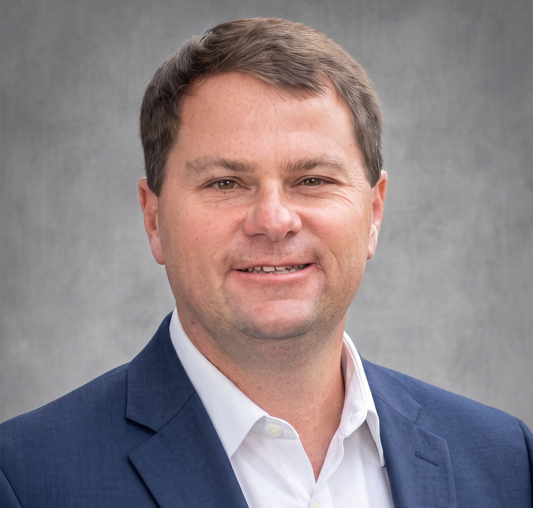 A man with short brown hair wearing a blue polo shirt stands in front of a plain gray background, looking at the camera with a neutral expression, as if ready to discuss topics such as water and soil management.
