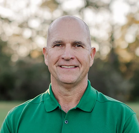 A middle-aged man with a shaved head is smiling outdoors, wearing a green collared shirt. The background is blurred with greenery and trees.