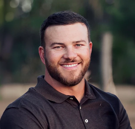 A man with short dark hair and a beard, wearing a dark polo shirt, smiles outdoors with a blurred green background.