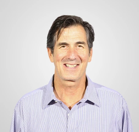 A middle-aged man with dark hair wearing a light purple striped shirt smiles at the camera against a plain light gray background.