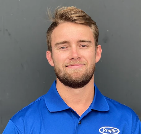 A man with light brown hair and a beard smiles slightly while wearing a blue collared shirt with a Profile logo, standing in front of a plain gray background.