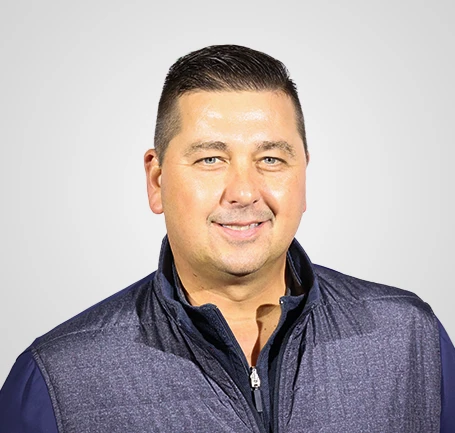 Smiling man with short dark hair wearing a navy blue zip-up vest over a collared shirt, standing in front of a plain light gray background.