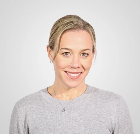 A woman with blonde hair pulled back, wearing a light gray sweater and a necklace, smiles at the camera against a plain light gray background.