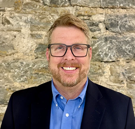 A man with light hair, glasses, and a beard, smiling and wearing a blue shirt and dark blazer, stands in front of a textured stone wall.