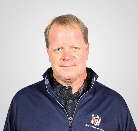 Middle-aged man with short blond hair wearing a navy blue pullover with an NFL logo, standing against a plain light gray background and looking at the camera.
