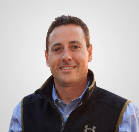 A man with short brown hair smiles at the camera. He is wearing a blue collared shirt and a black Under Armour vest. The background is light gray and plain.