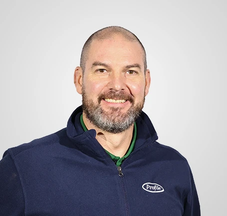 A smiling man with a beard, wearing a navy blue jacket with a Profile logo, stands against a plain light gray background.