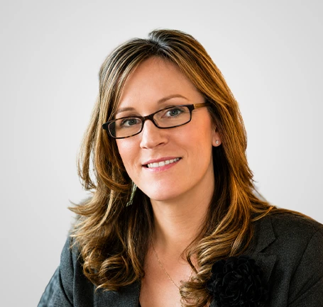 Woman with long, wavy brown hair and glasses, wearing a gray blazer and smiling, posed against a plain light background.