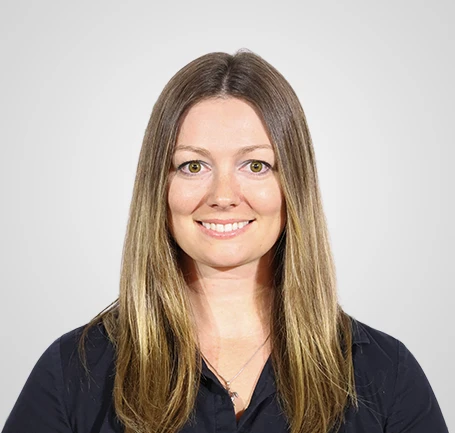 A woman with straight, long brown hair and green eyes, smiling at the camera. She is wearing a dark shirt and a necklace, with a plain light gray background.