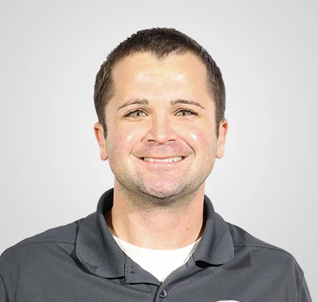 A man with short brown hair smiles at the camera. He is wearing a dark gray collared shirt over a white undershirt, with a plain light gray background behind him.