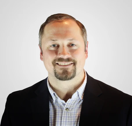 Smiling man with short hair, a trimmed beard, and mustache, wearing a dark jacket over a light checkered shirt, in front of a plain light background.