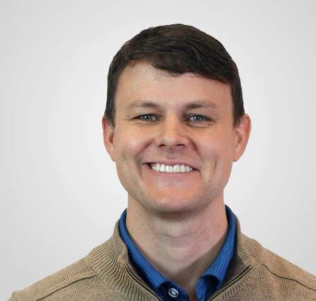Smiling man with short brown hair wearing a tan sweater over a blue collared shirt, pictured against a plain light gray background.