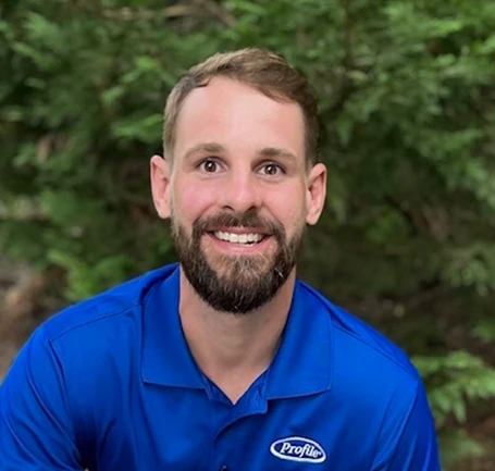 A bearded man wearing a blue collared shirt with a Profile logo smiles at the camera, sitting outdoors with green trees in the blurred background.