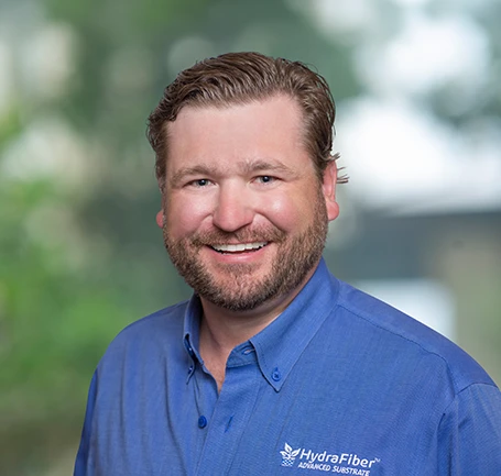 Smiling man with short, light brown hair and a beard, wearing a blue HydraFiber shirt, posed in front of a blurred outdoor background.